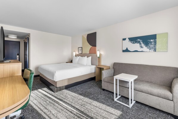 Habitación de hotel con cama, sofá, una mesita, escritorio y un cuadro en la pared.