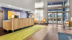 Un espace d’accueil avec un bureau en bois, deux chaises orange, un tapis vert et de grandes fenêtres donnant sur l’extérieur.