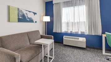 Une pièce avec un canapé gris, une petite table blanche, un lampadaire, une fenêtre avec des rideaux légers et un climatiseur fixé au mur.