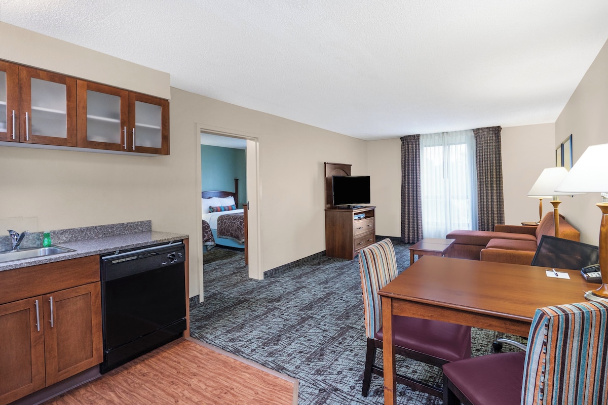 Habitación de hotel con kitchenette, zona de comedor, sala de estar con sofá y televisor, y baño visible a través de una puerta abierta.