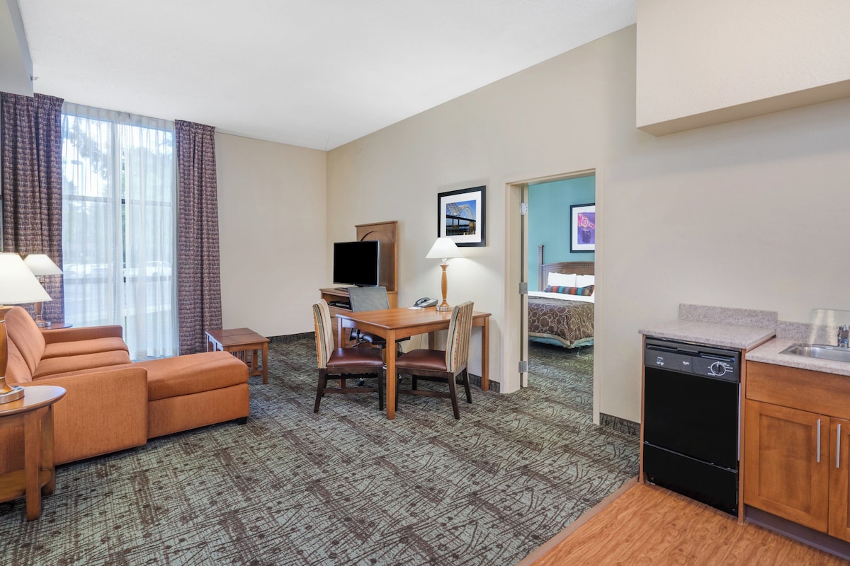 Habitación de hotel con sofá, una mesa de comedor con sillas, televisor, cama y una zona de cocina.