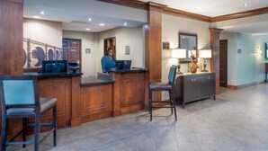 A reception area with a wooden reception desk, a computer, and a telephone. There are two chairs and a cabinet with a mirror and a vase of flowers.
