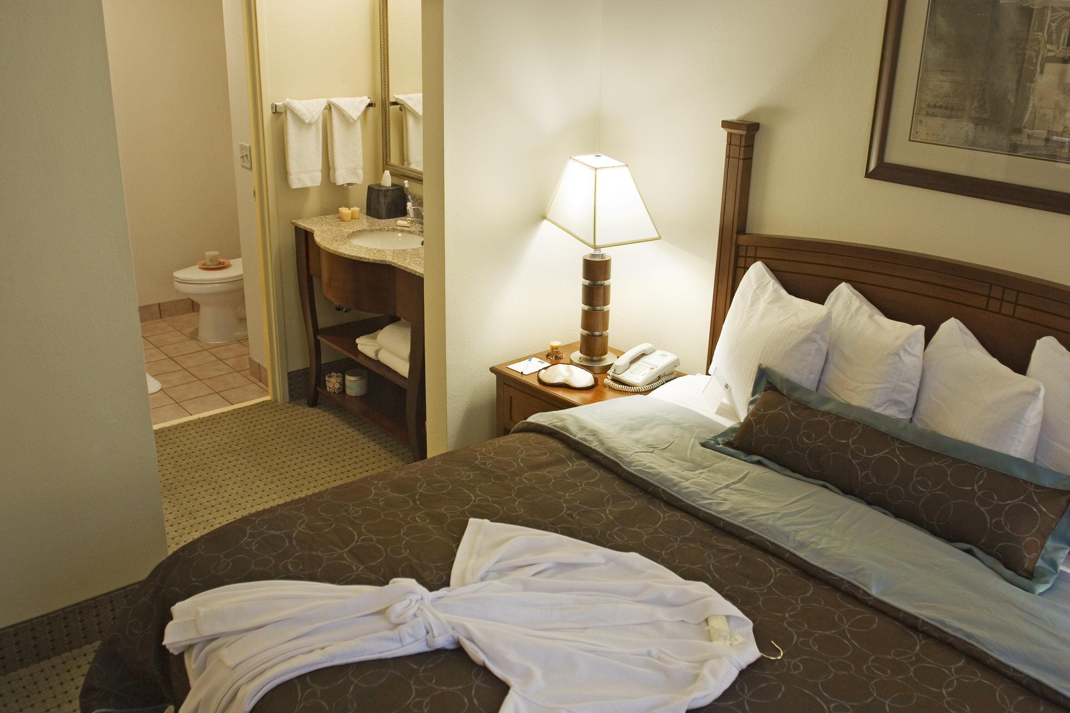 A hotel room with a bed, a bedside table with a lamp, a phone, and a bathroom visible through an open door.