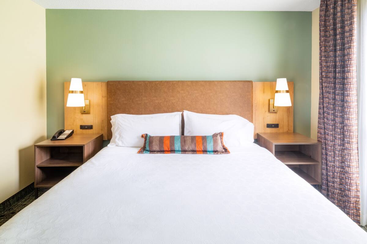 Habitación de hotel con una cama grande, dos mesitas de noche con lámparas, un teléfono y un cojín decorativo estampado.