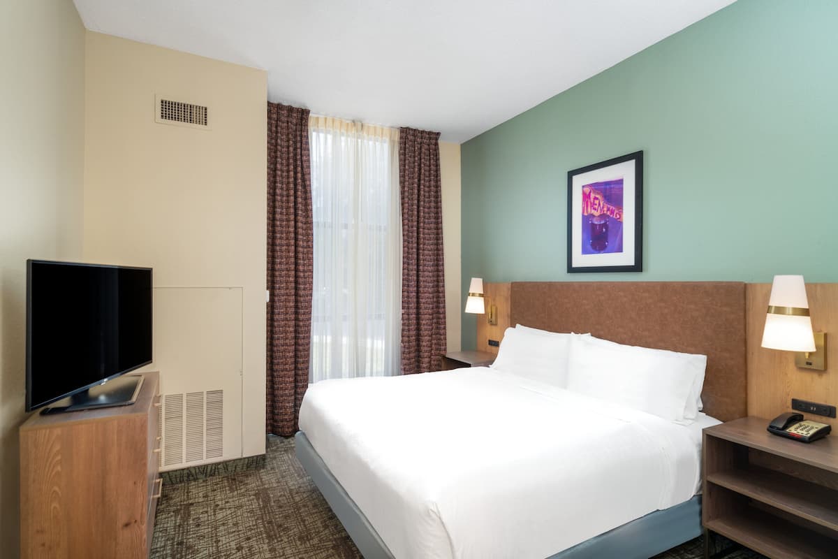 Una habitación de hotel con una cama, un televisor sobre un mueble de madera, una mesita de noche con una lámpara y un cuadro en la pared.