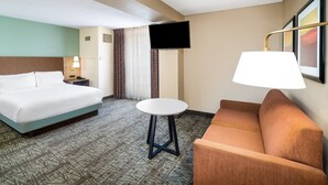 A hotel room with a bed, a sofa, a small table, a TV mounted on the wall, and a window with curtains.