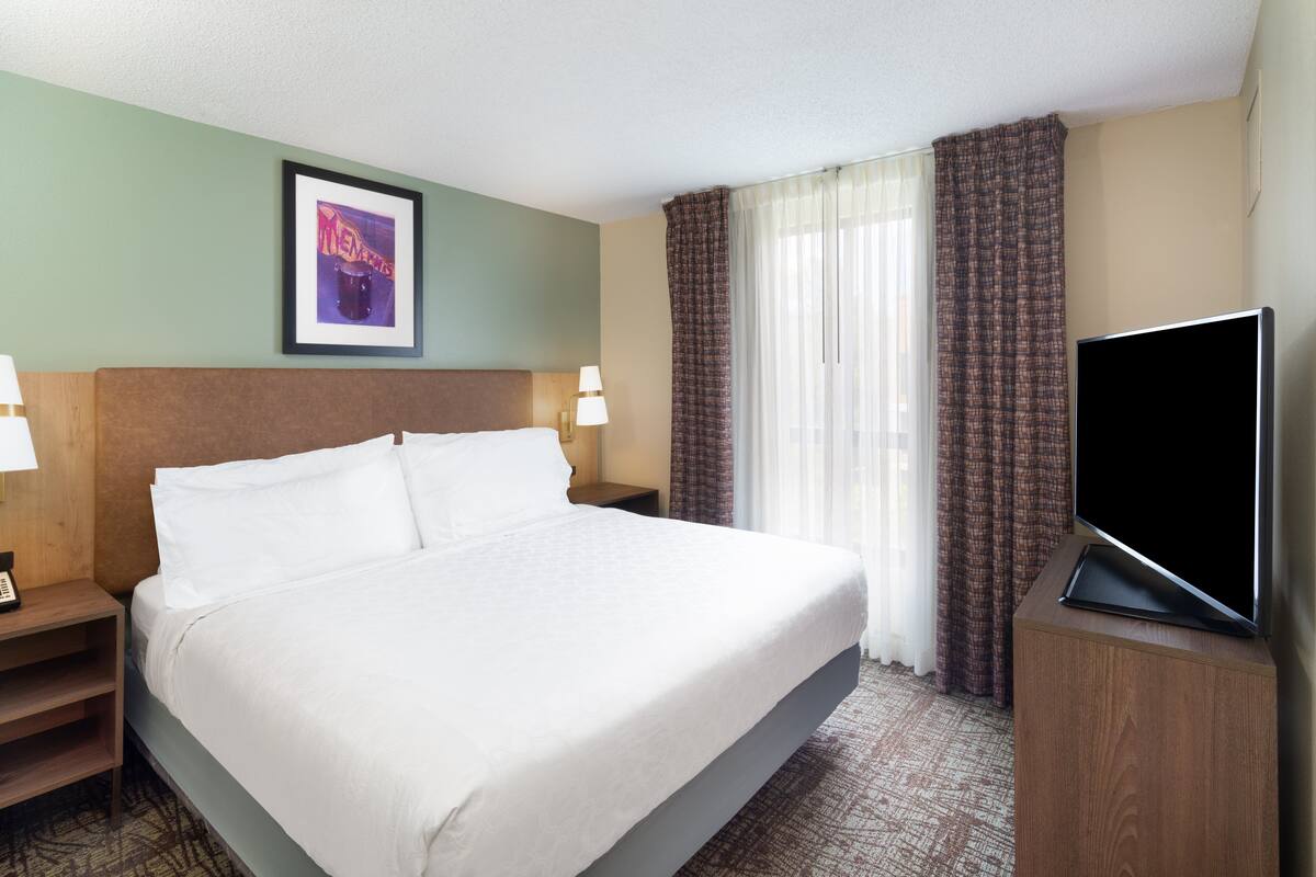 Habitación de hotel con una cama grande, televisor de pantalla plana, mesita de noche con lámpara y un cuadro en la pared.