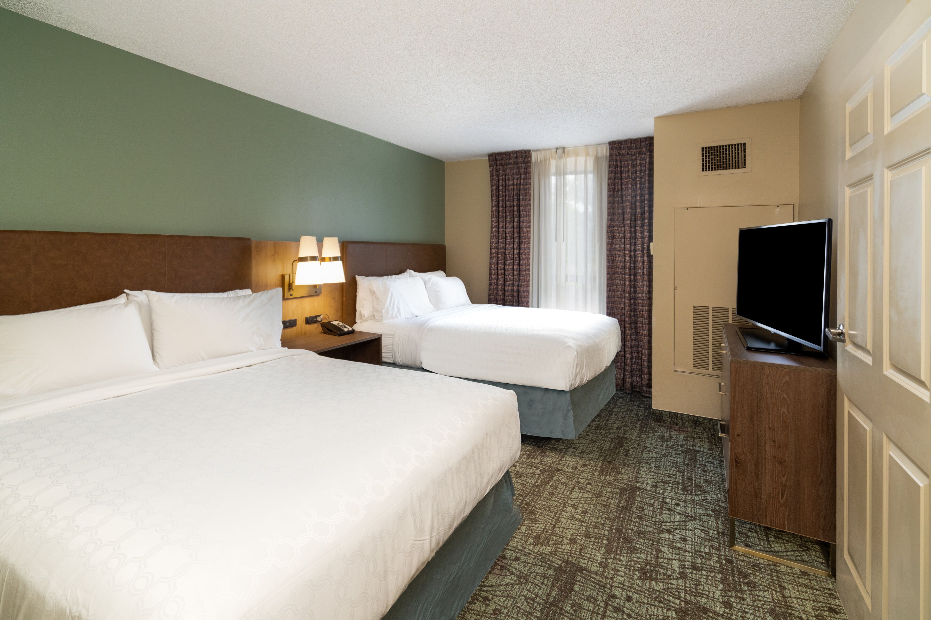 Quarto de hotel com duas camas, televisão, escrivaninha com abajur e janela com cortinas.