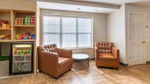 Una habitación con dos sillones de cuero marrón, una mesita redonda pequeña, un refrigerador lleno de bebidas y un ventanal grande con persianas.