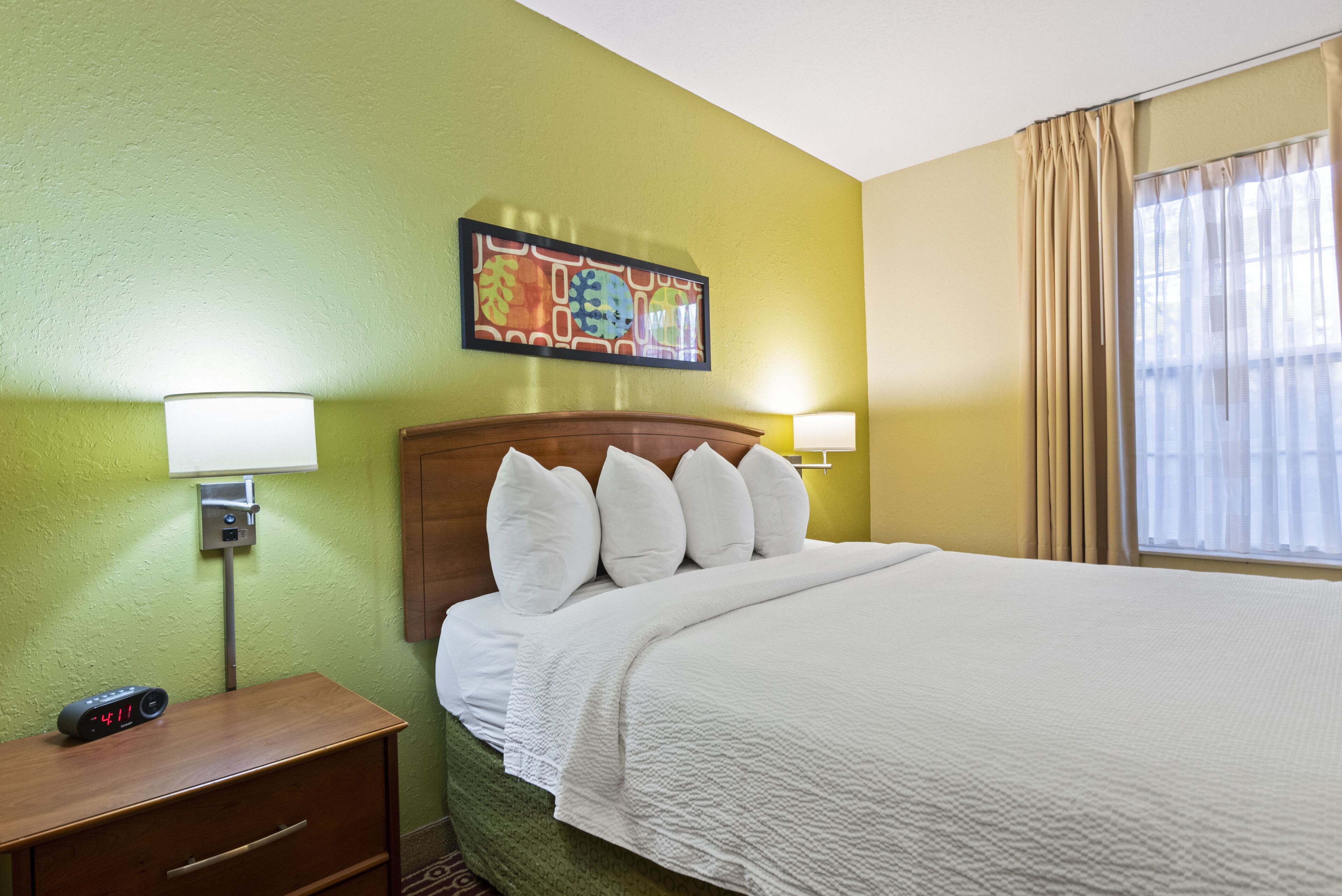 A hotel room with a bed, bedside lamps, a nightstand, a clock, and a painting on the wall.