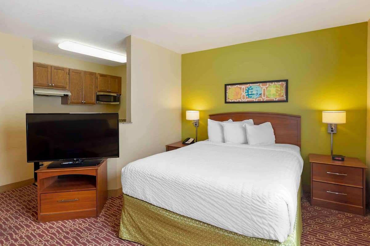 Une chambre d’hôtel avec un lit, des lampes de chevet, un téléviseur posé sur un meuble en bois, un petit tiroir, un four à micro-ondes et un tableau accroché au mur.