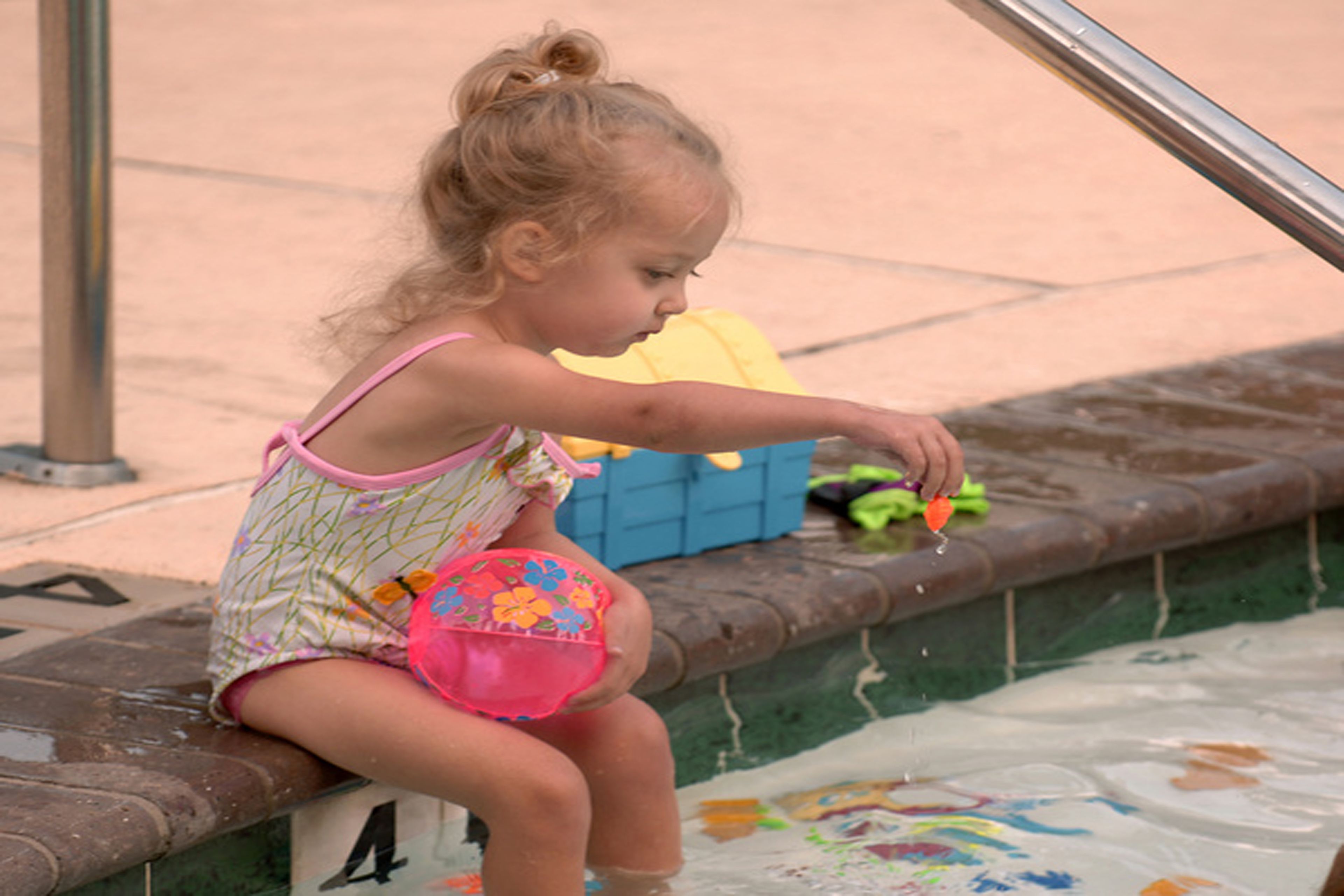 A child sitting by a pool with a numbered bench, a yellow toy, and a pink bucket.
