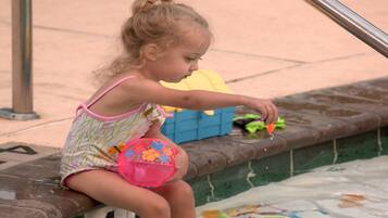 A child sitting by a pool with a numbered bench, a yellow toy, and a pink bucket.