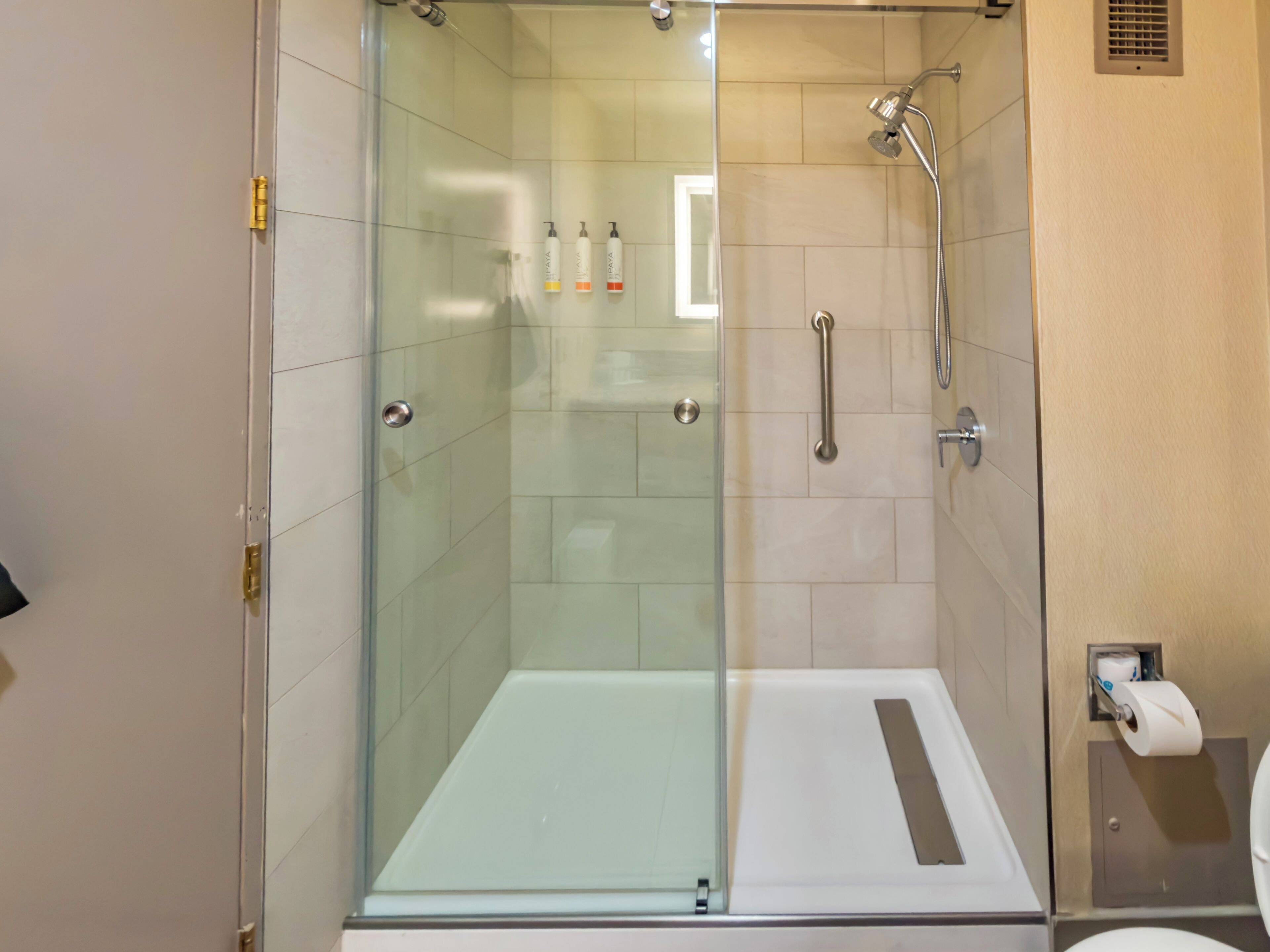 A glass-enclosed shower stall with a white floor, a handheld showerhead, and a toilet paper dispenser on the wall.