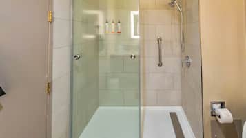 A glass-enclosed shower stall with a white floor, a handheld showerhead, and a toilet paper dispenser on the wall.