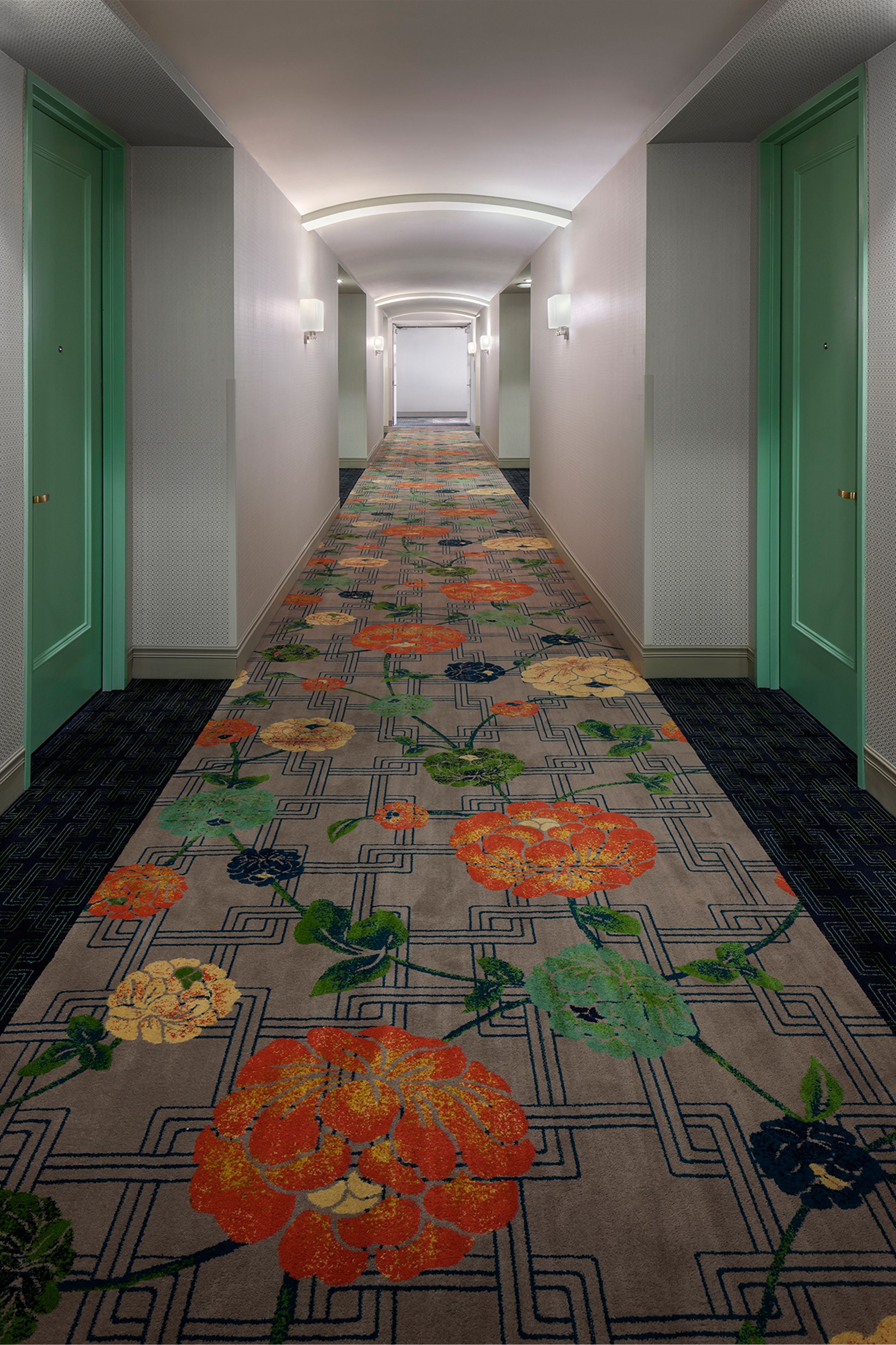 Un pasillo de hotel con puertas verdes, un suelo estampado con flores y un largo corredor que termina con una lámpara.