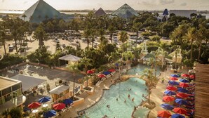 A resort with a large swimming pool surrounded by palm trees and umbrellas, a parking area, and a view of pyramids in the background.