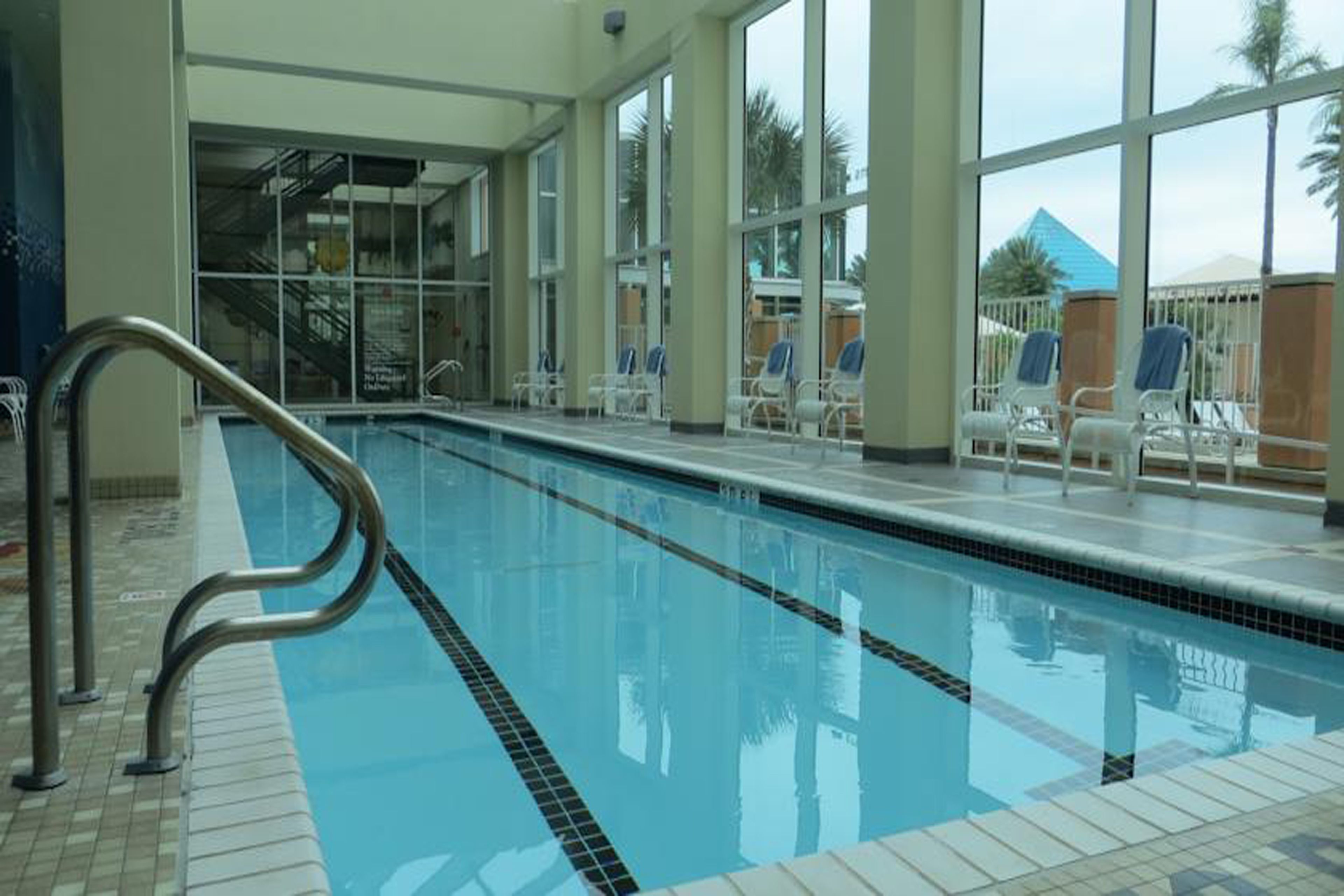 En indendørs swimmingpool med glasvægge, hvide liggestole og flisegulv.