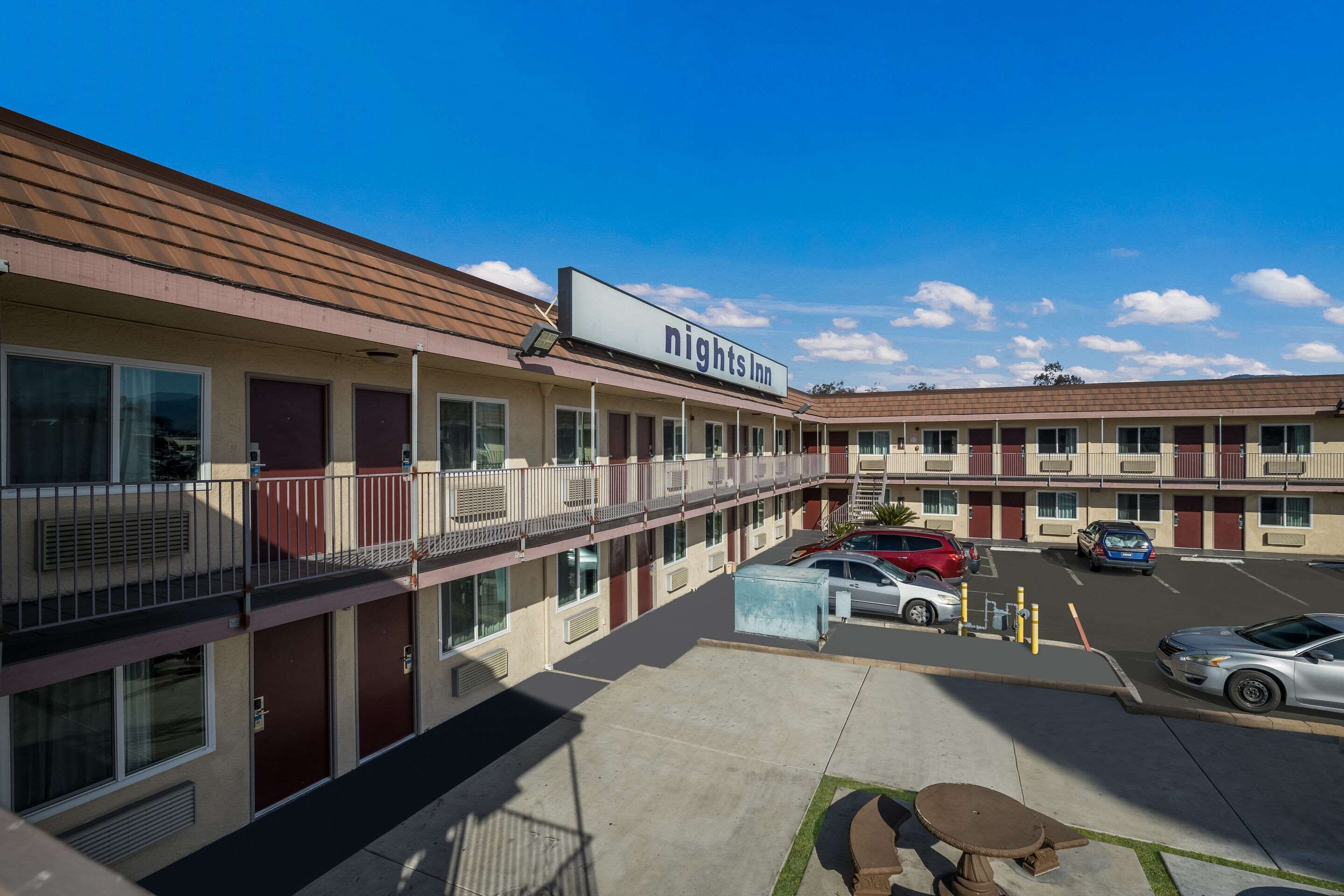 Een motel met een bord waarop 'Nights Inn' staat, bestaande uit een gebouw met twee verdiepingen, balkons, een parkeerplaats en een helderblauwe lucht.