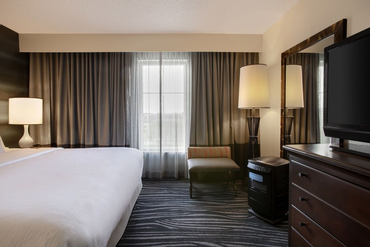 Habitación de hotel con cama, escritorio, televisor y ventana con cortinas.