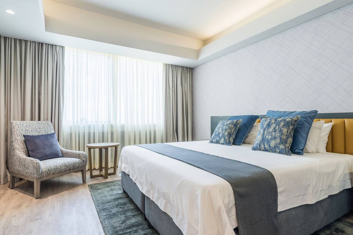 Ein Hotelzimmer mit einem großen Bett, einem Sessel, einem kleinen Tisch und einem Fenster mit Vorhängen.