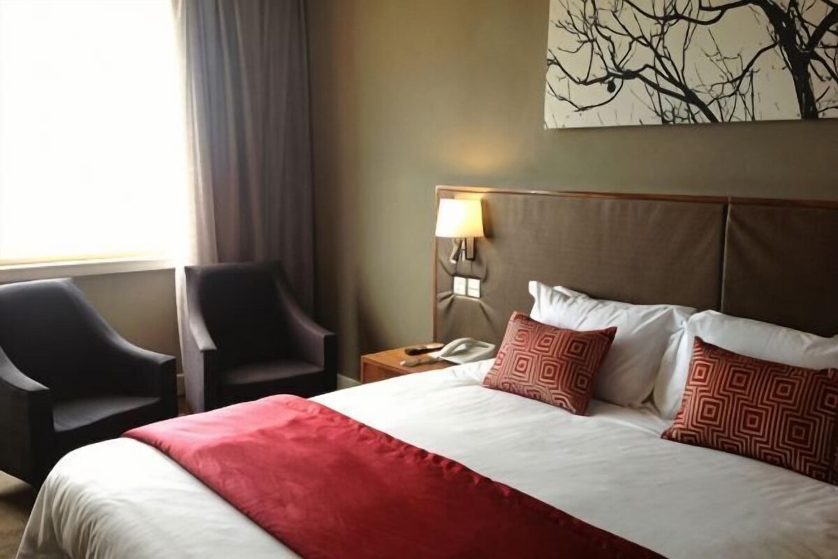 A hotel room with a bed, two chairs, a nightstand, a lamp, and a wall-mounted artwork.