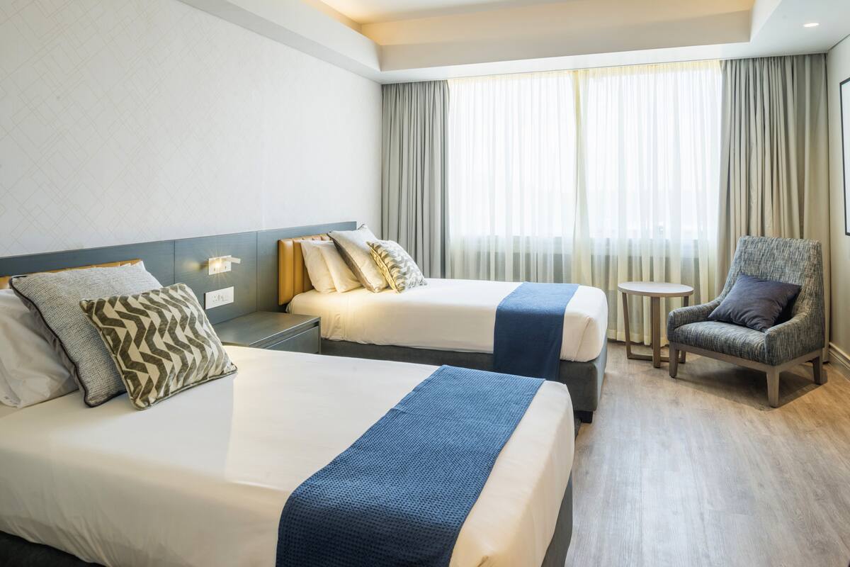 Ein Hotelzimmer mit zwei Betten, einem Sessel, einem kleinen Tisch und einem Fenster mit Vorhängen.