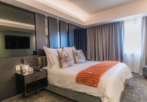 Habitación de hotel con una cama grande, una mesita de noche con un teléfono y una botella, un televisor de pantalla plana montado en la pared y una ventana con cortinas.