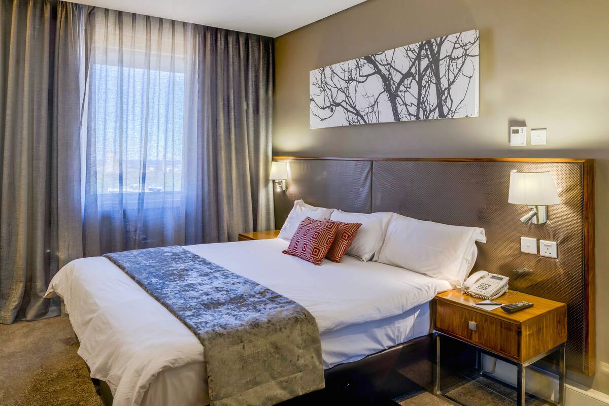 Ein Hotelzimmer mit einem großen Bett, einem Nachttisch mit Telefon, einer Lampe und einer Wandleuchte. Es gibt ein Fenster mit Vorhängen und ein Bild mit einem Ast als Motiv an der Wand.