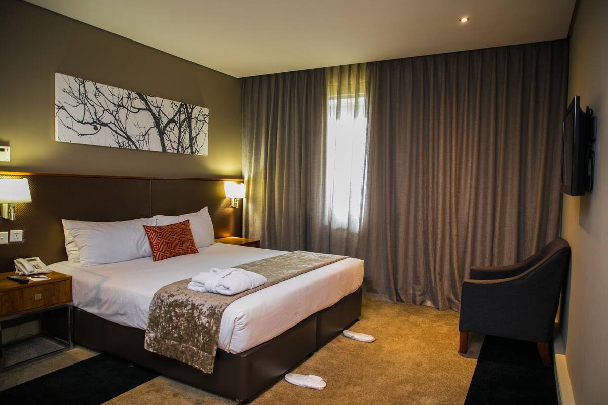 Ein Hotelzimmer mit einem großen Bett, einem Schreibtisch mit Telefon, einem Sessel und einem an der Wand befestigten Fernseher.