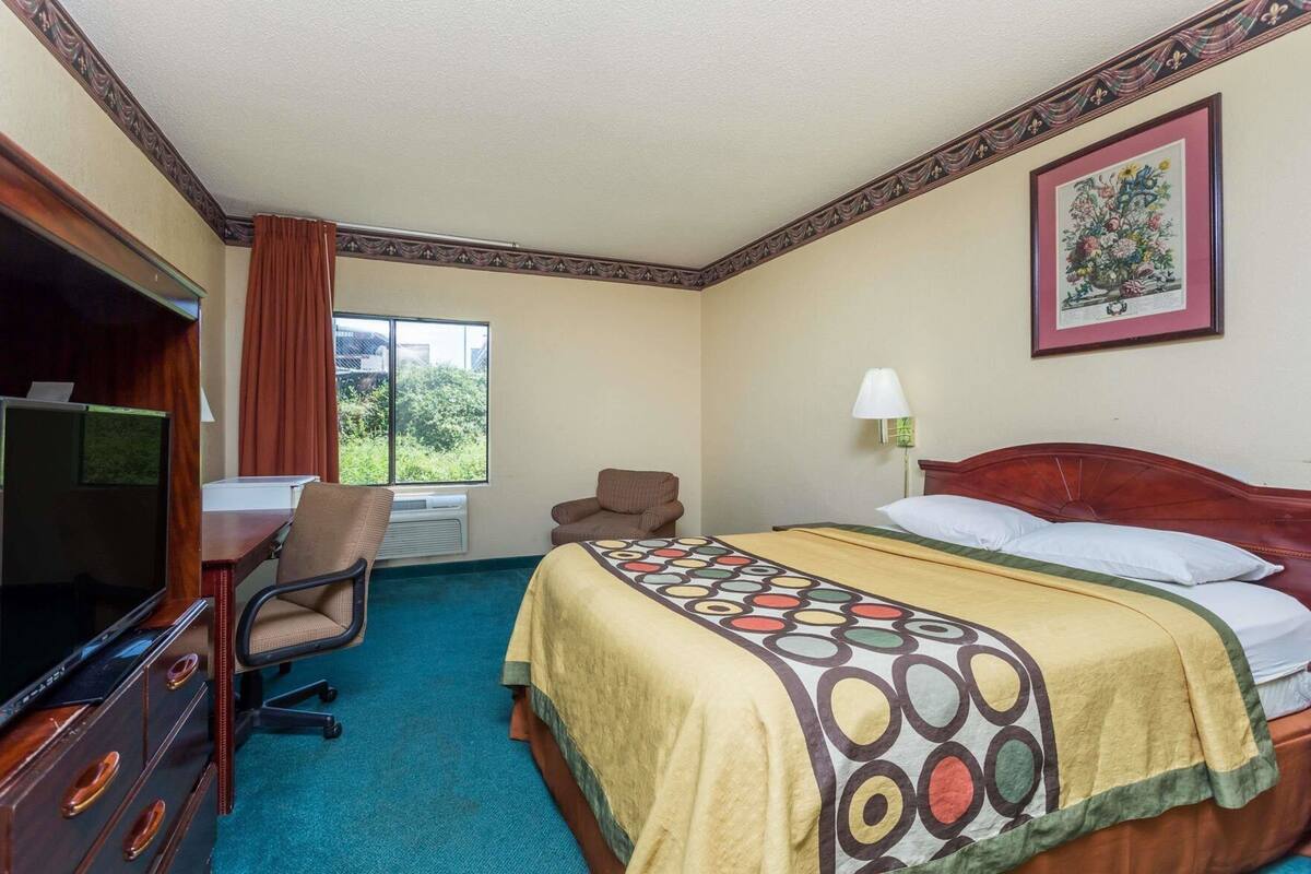 Quarto de hotel com cama, escrivaninha com cadeira, televisão e janela com cortinas.