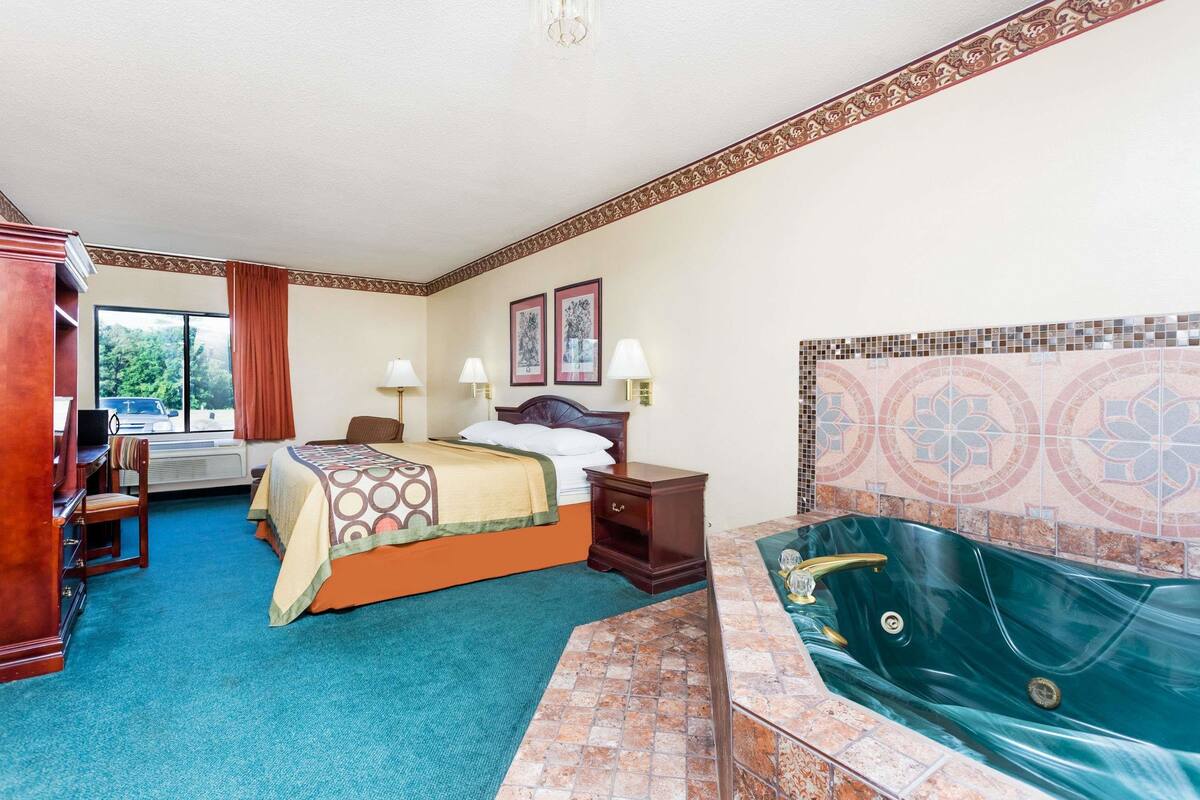 Quarto de hotel com cama, escrivaninha, cadeira, abajur, mesa de cabeceira, TV fixada na parede, janela com cortinas e banheira com parede em mosaico.