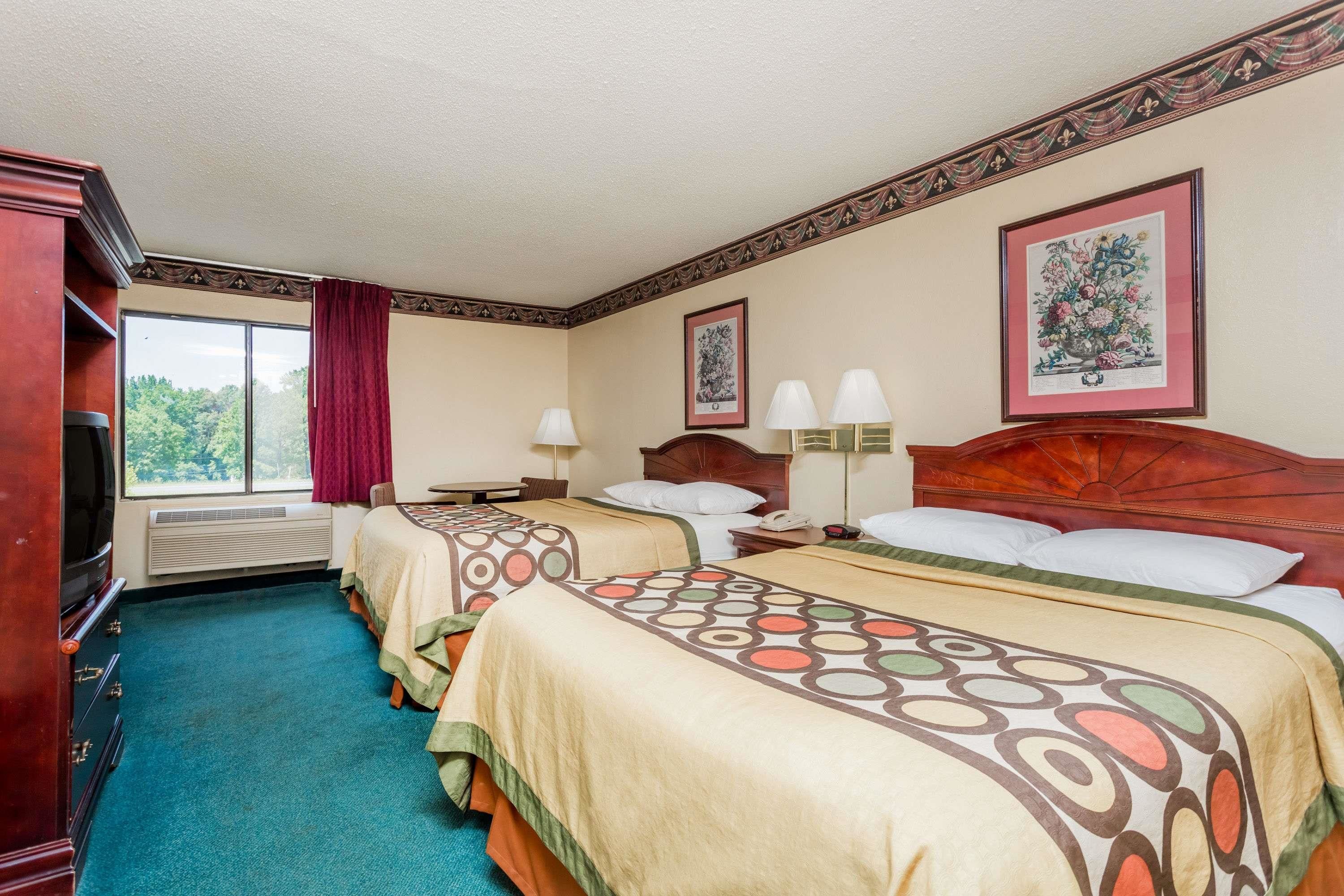 A hotel room with two beds, a large window with a view of trees, a wooden headboard, and a framed botanical print on the wall.