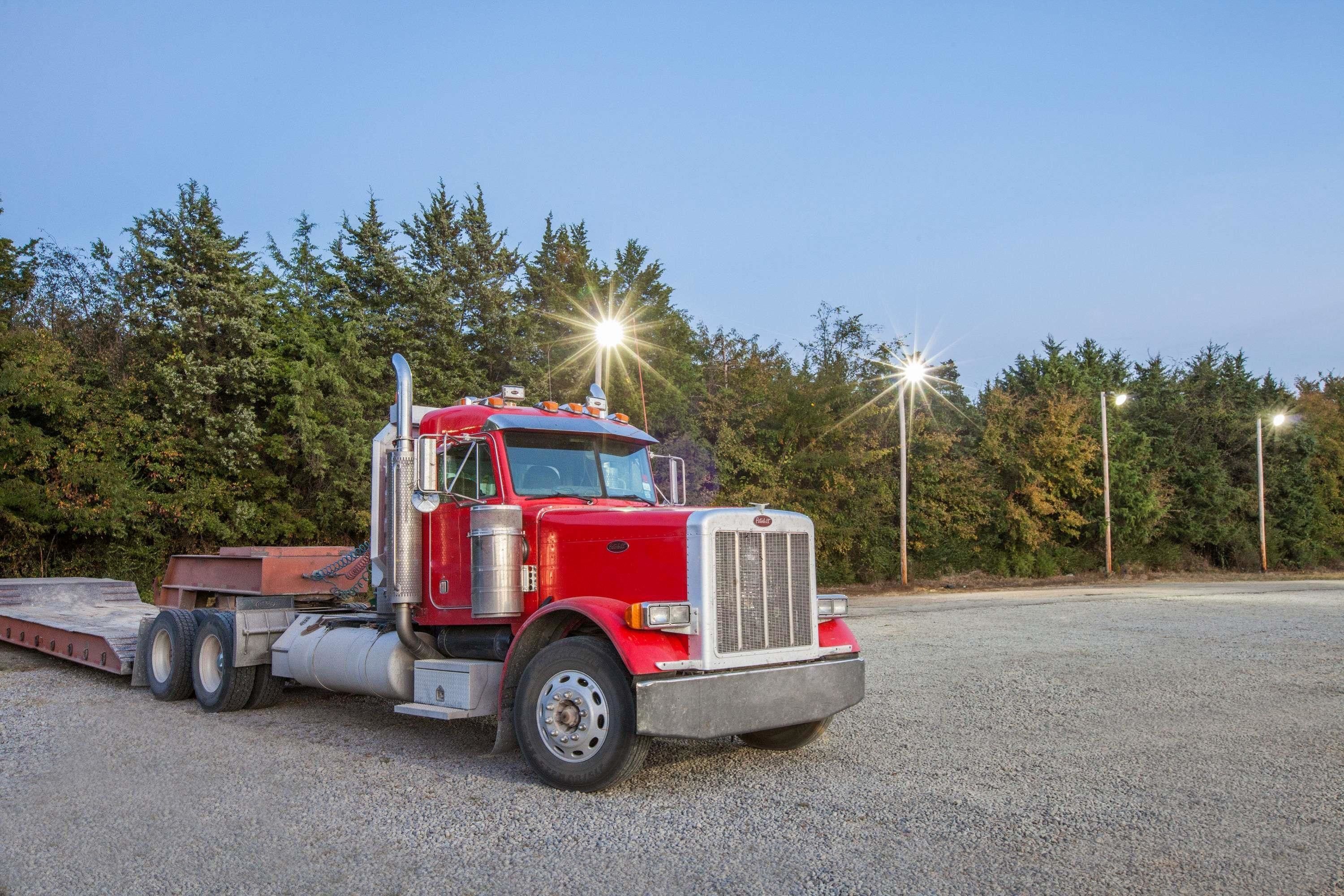 Un camion semi-remorque rouge est garé sur une surface de gravier, avec une remorque attelée, et il est entouré d’arbres et de lampadaires.