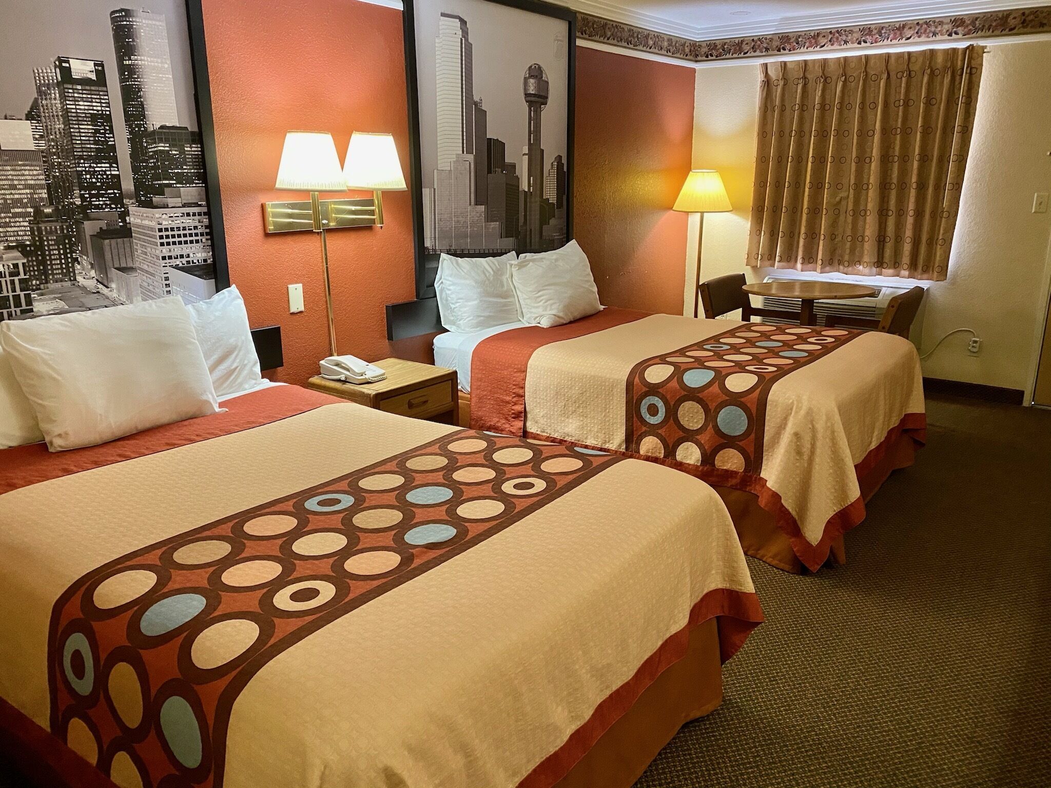 Habitación de hotel con dos camas, un mural de paisaje urbano, una lámpara y una ventana con cortinas.