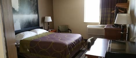 Habitación de hotel con cama, escritorio, silla, lámpara, ventana con cortinas, aire acondicionado y un cuadro en la pared.