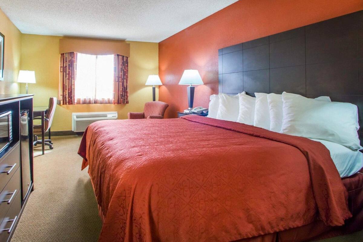 A hotel room with a large bed, two bedside lamps, a desk with a chair, and a window with curtains.