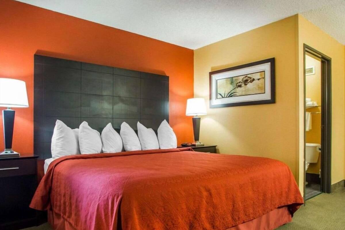 A hotel room with a bed, a nightstand, a lamp, a framed picture on the wall, and a door leading to a bathroom.