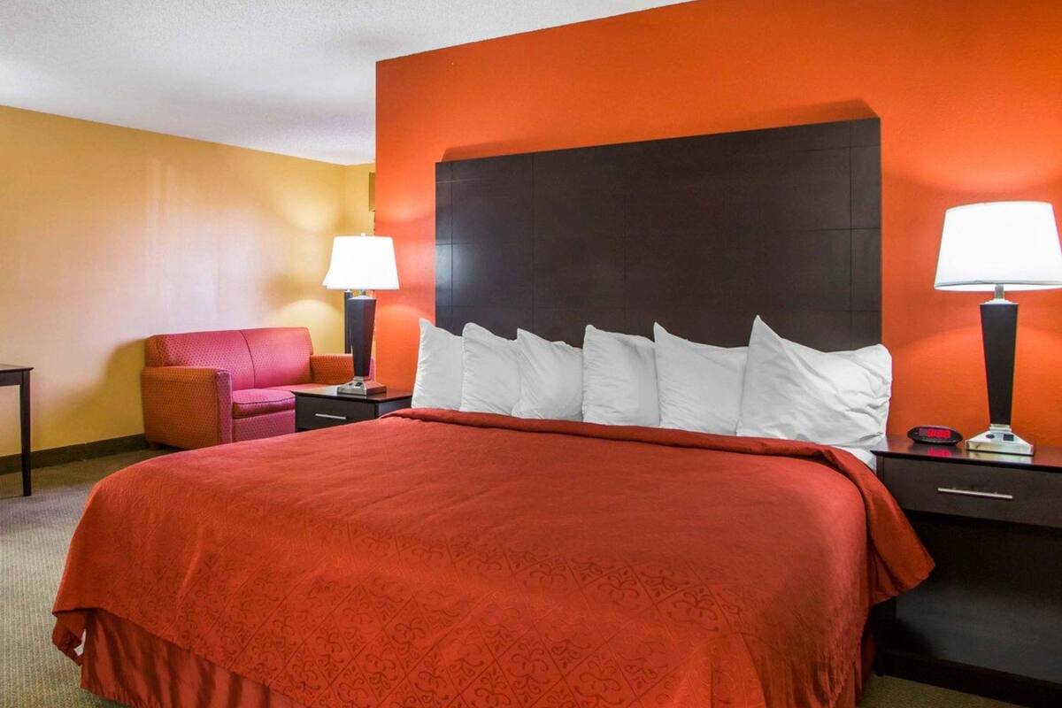 A hotel room with a large bed, a red upholstered chair, two bedside tables with lamps, and a dark headboard.
