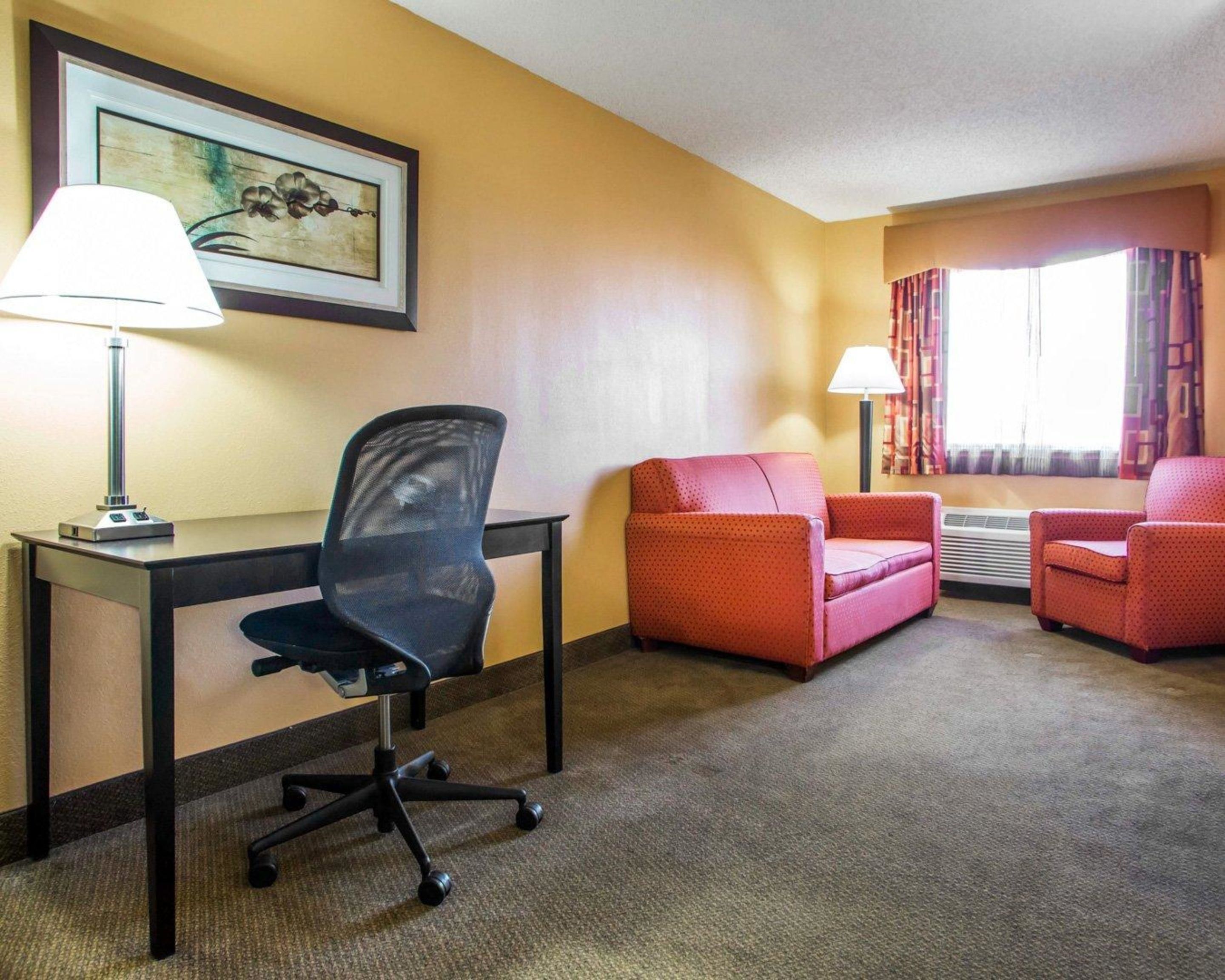 A hotel room with a desk, two chairs, a lamp, a painting, and a window with curtains.