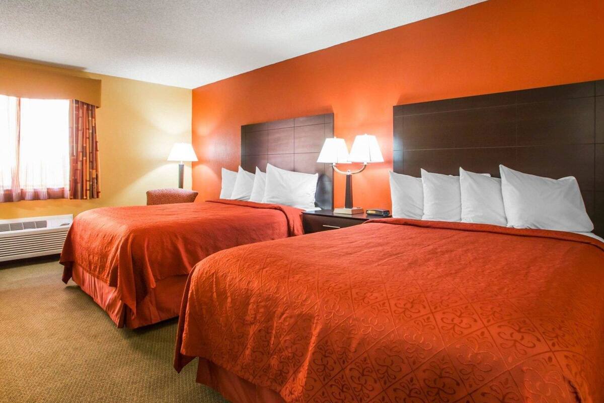 A hotel room with two beds, each with a red bedspread and white pillows, a nightstand with a lamp, and a window with curtains.