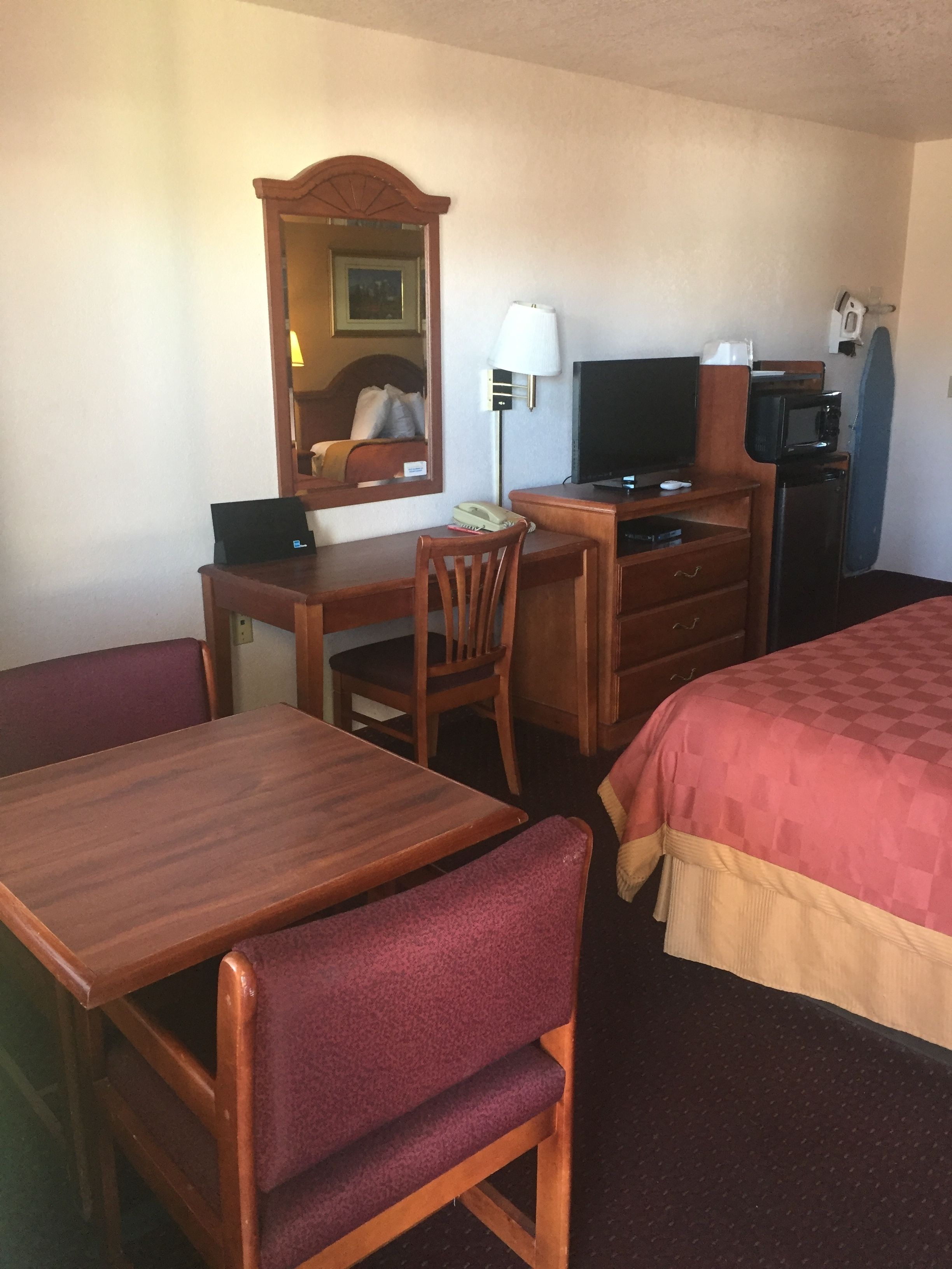 Een hotelkamer met een houten bureau, een stoel, een bed met ruitbeddengoed, een televisie, een magnetron en een spiegel.