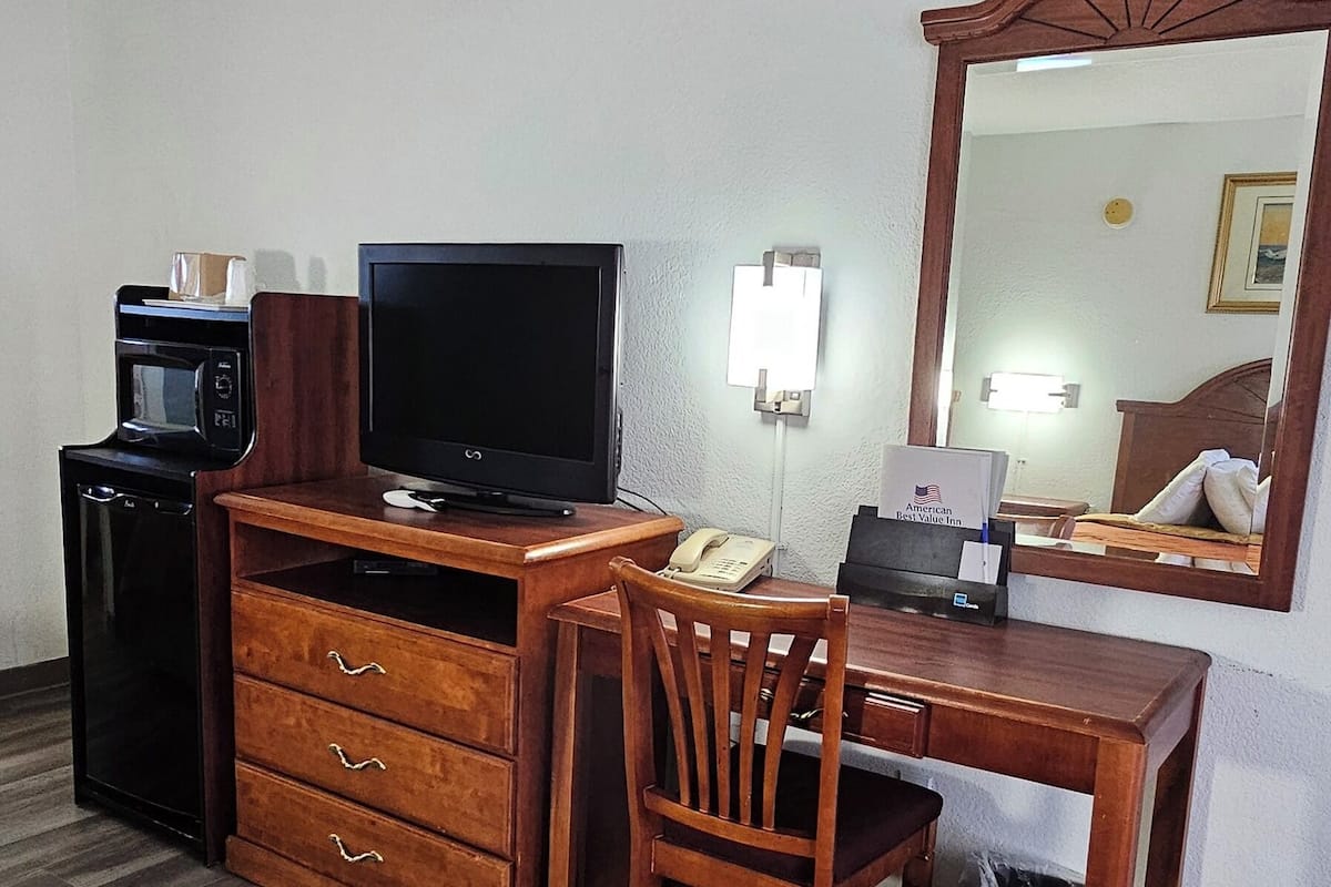A hotel room with a wooden desk, a television, a microwave, a trash bin, and a bed with an orange blanket.