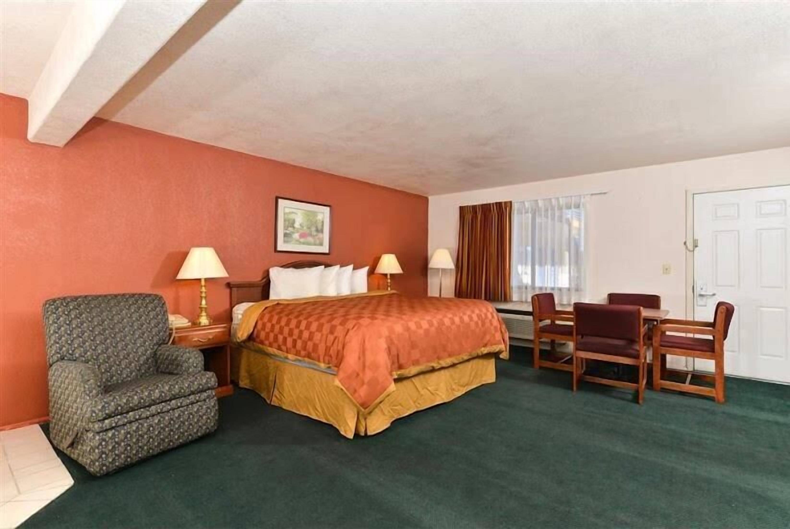 A hotel room with a bed, two bedside tables with lamps, a chair, a dining table with chairs, and a window with curtains.