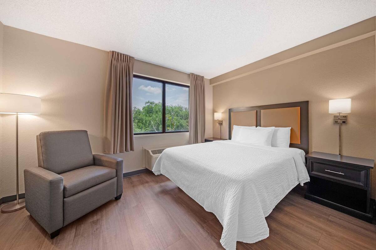 A hotel room with a bed, a chair, a nightstand, a lamp, and a window with curtains.