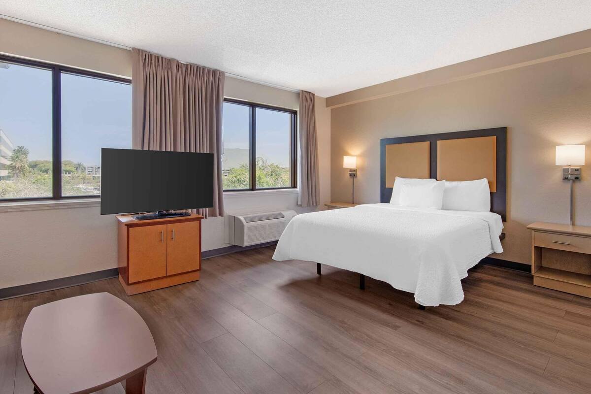 A hotel room with a bed, a TV on a wooden cabinet, a bedside table with a lamp, and a view of the outdoors through large windows.