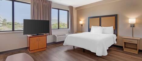 A hotel room with a bed, a TV on a wooden cabinet, a bedside table with a lamp, and a view of the outdoors through large windows.