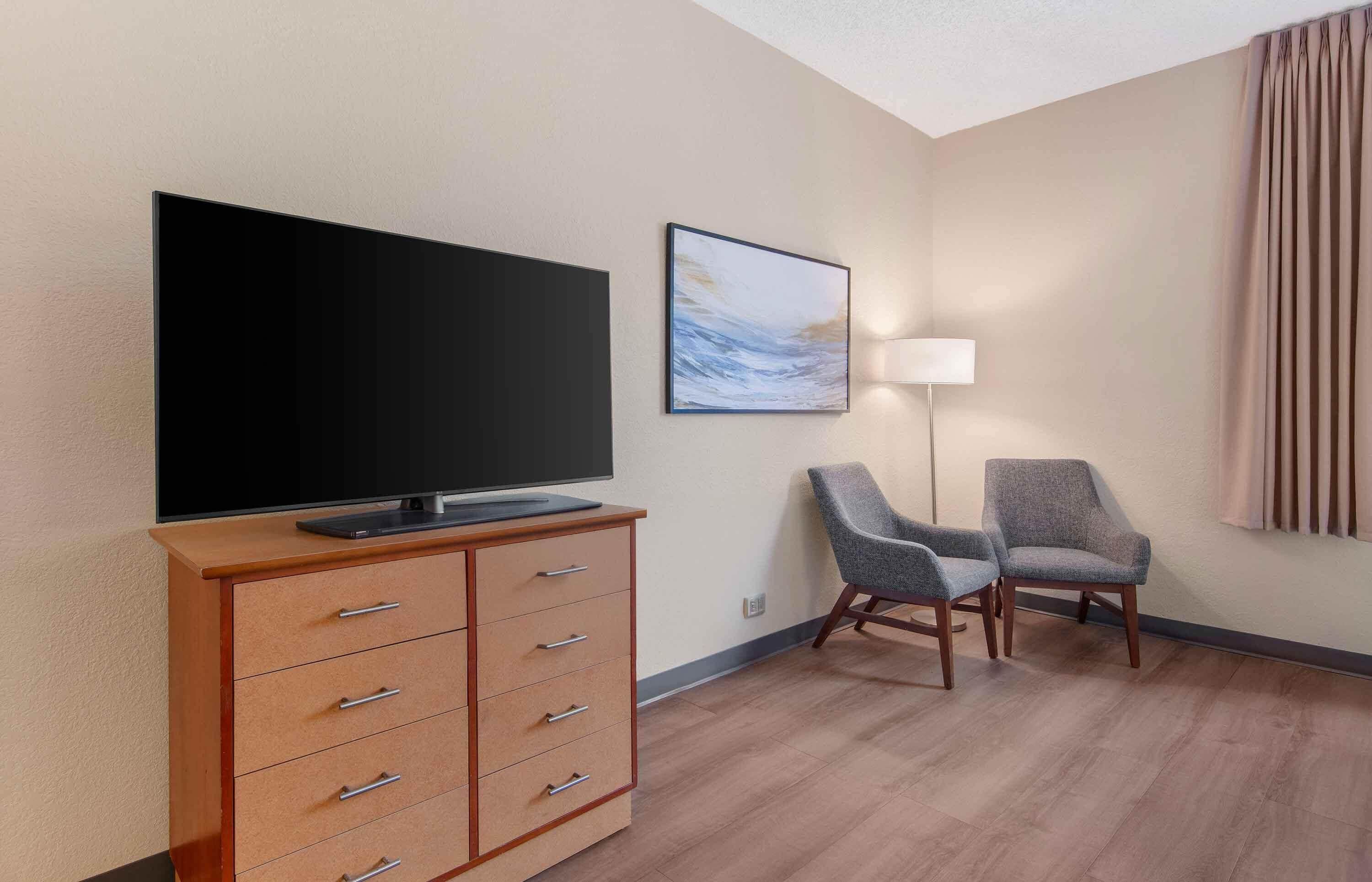 A hotel room with a flat-screen TV on a wooden cabinet, two grey armchairs, a floor lamp, and a painting on the wall.