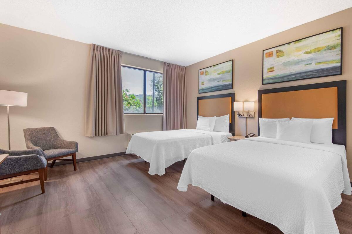 A hotel room with two beds, a chair, a lamp, and a painting on the wall.