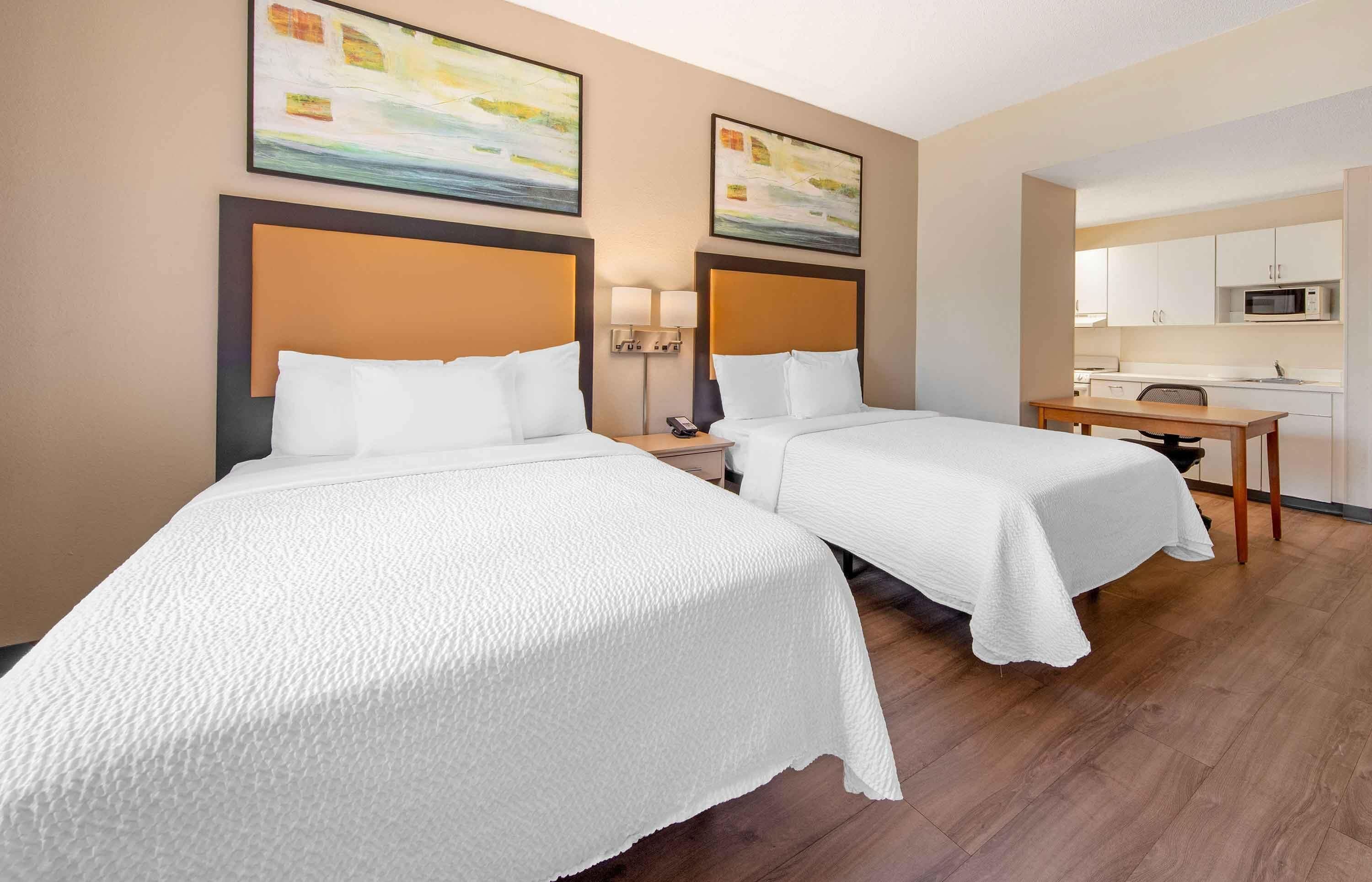 A hotel room with two beds, a desk, a chair, a microwave, and a small kitchen area.